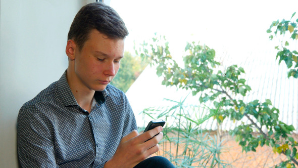 Teenager Sitting By A Window Using His Smartphone