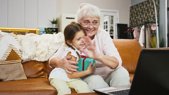 Grandma Smiling and Hugging Grandchild Holding Gift Box Saying Hi to Someone Using Online Video Call