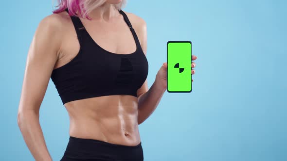 Sporty Woman Holds Phone with Green Screen Pointing at It with Her Finger on Blue Background Closeup