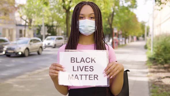 A Young Black Woman in a Face Mask Shows a Black Lives Matter Sign To the Camera in the Street