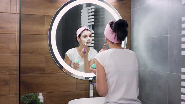 Young Woman with Headband Cleaning Her Face with Cosmetic Foam, Standing in Front of Mirror