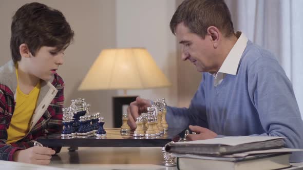 Side View Close-up of Cute Little Caucasian Boy Playing Chess with Grandfather Indoors. Grandson