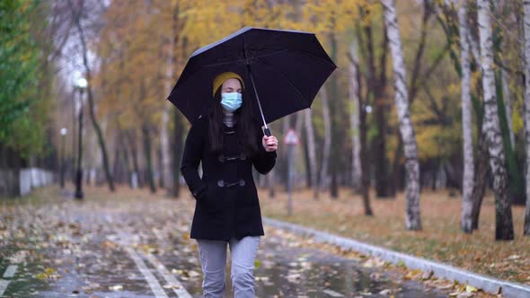 A Young Woman in a Protective Mask Walking in the Park Under Umbrella. Rainy Day, During Second Wave