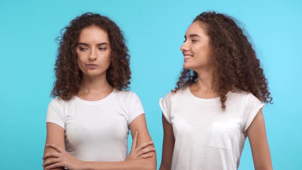 One Dissapointed Female Curly Caucasian Twin Standing on Blue Background While Another Smiling and