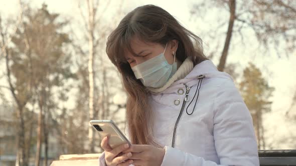 Young Woman in a Medical Protective Mask Is Sitting with a Smartphone on City Street in Europe