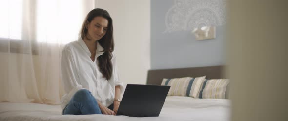 A young woman smiling while working on a laptop from her bedroom. 