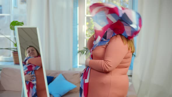 Joyful Fat Female in Dress and Bright Scarf Admiring Her Mirror Reflection Standing in Living Room