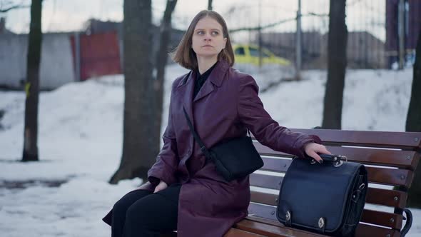 Worried Caucasian Woman in Purple Coat Sitting on Bench in Snowy Park Sighing Putting Handbag on