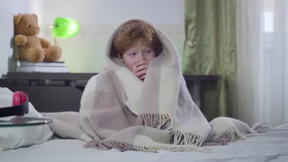 Portrait of Cute Little Caucasian Boy Sitting on Bed Covered with Blanket and Looking Around. Scared