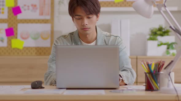 Asian young man sits at his desk and begins turn on and use his laptop for work from his home