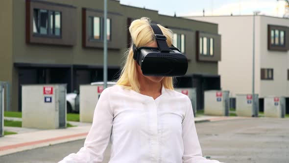 Young Attractive Blond Woman Uses Virtual Reality Glasses - Buildings in the Background