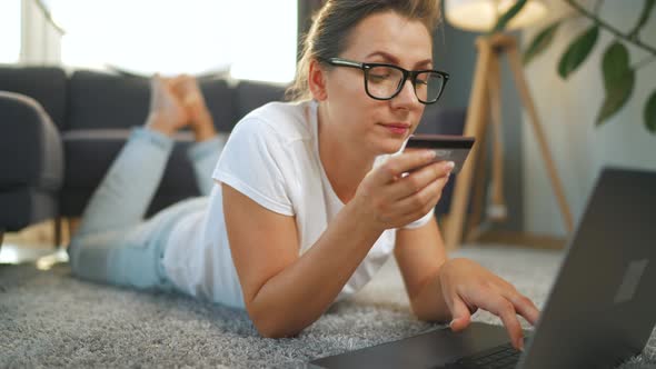 Woman with Glasses is Lies on the Floor and Makes an Online Purchase Using a Credit Card and Laptop