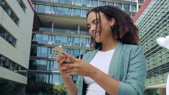 Happy Satisfied Mixed Race Young Woman with Curly Hair Stylishly Dressed Using Her Smartphone While