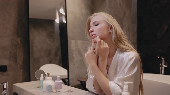 Young Blonde Woman in a White Robe in the Bathroom with a Massage Stone