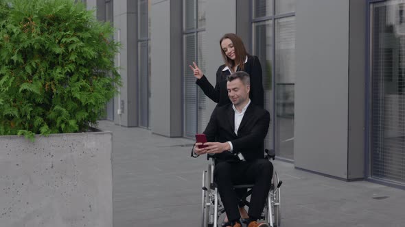 Cheerful Woman and Man in Wheelchair Posing To Smartphone Camera While Standing Near Office Building