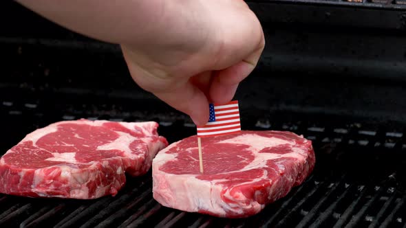 Two juicy rib-eye steaks sitting on the grill and cooking a hand comes in and sticks a tiny American