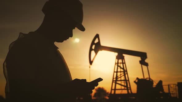 Silhouette of Man Engineer with Phone Overseeing the Site of Crude Oil Production at Sunset.