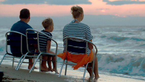 Family Of Three Sitting On Chairs By Sea At Sunset