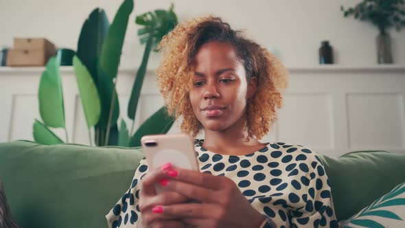 Young African American Woman Chats in Phone Sits on Sofa in Living Room