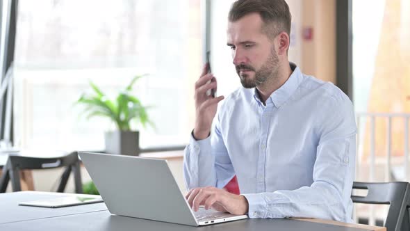 Young Man with Laptop Talking on Smartphone in Office 