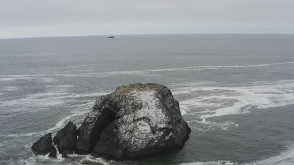 Stormy day aerial of a rocky ocean shore, panning around a large rock in the ocean, waves crash agai