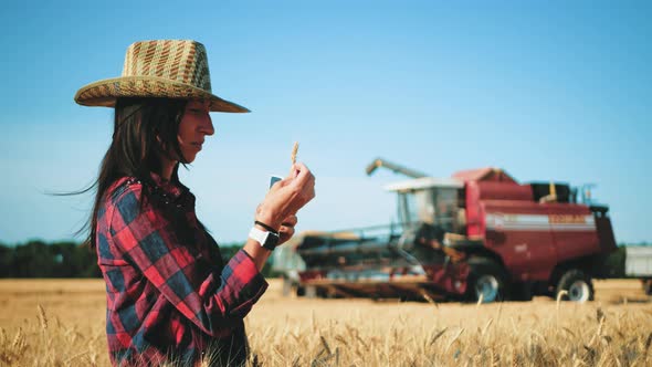 Young Woman Farmer in Hat Standing Against the Background of a Working Combine Harvester at Sunset