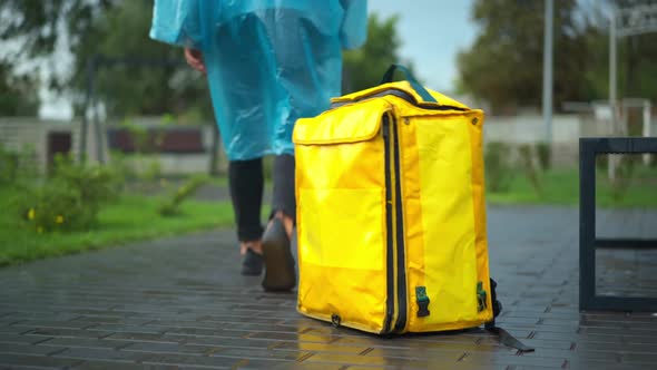 Unrecognizable Young Man in Rain Coat Leaving Yellow Delivery Backpack Walking Away in City Park