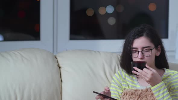 Girl at Home on the Sofa in the Evening Drinks Tea and Uses the Phone