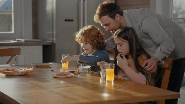 Man Hugs His Children Drinking Orange Juice at Breakfast on a Sunny Day