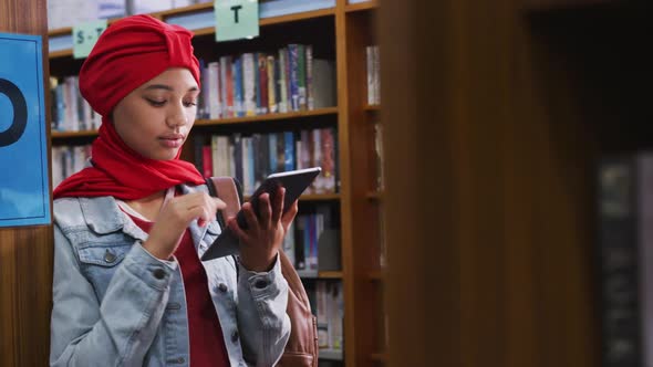 An Asian female student wearing a red hijab studying in a library and using tablet