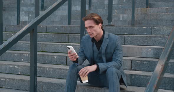 Young Handsome Businessman with Coffee in His Hands Sits on the Steps Near the Office Building and