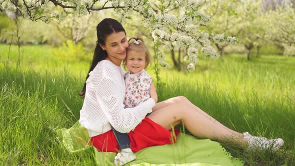 Mom with a Daughter at a Picnic on the Lawn in the Park By a Flowering Tree