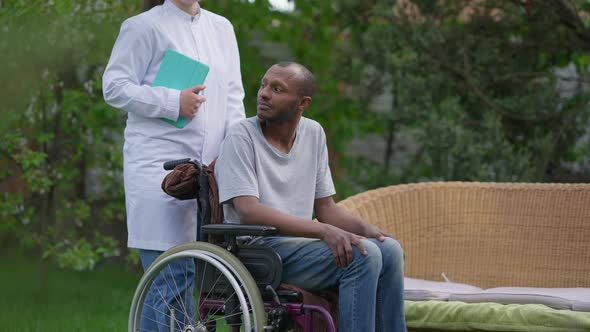 Thoughtful Crippled African American Man in Wheelchair Looking Away Talking to Unrecognizable