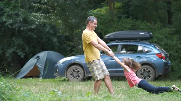 Happy father spinning his smiling daughter child around holding her hands at camping site in summer.
