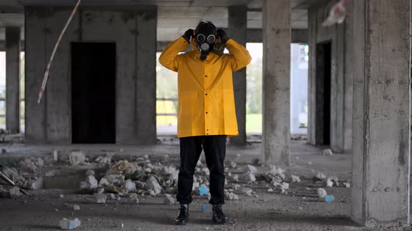 Stalker Survivor Stands in Yellow Raincoat and Black Gas Mask in Abandoned Building and Wears Hood