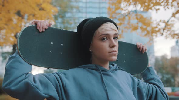 Young Blond Caucasian Woman Skater Holding Skateboard Behind Her Head in the Park in Autumn