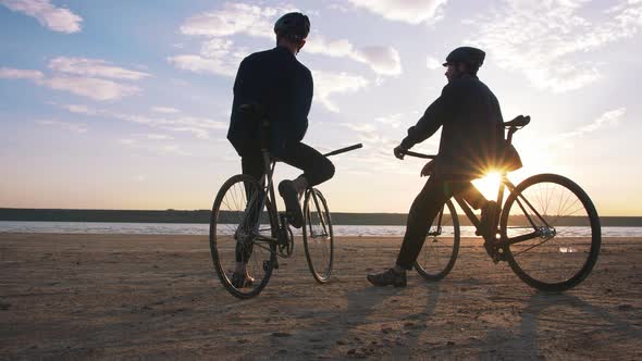Two Young Men Sitting on Bicycles on the Beach on the Background of an Orange Sunsetting Sky