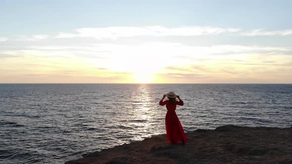 Aerial View of a Young Woman in a Red Dress and a Straw Hat Walks Along the Edge Front of a Cliff By