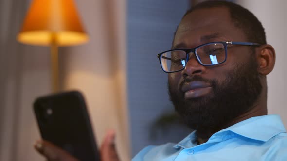 Smiling African American Man Holding Smartphone Using Social Media Apps Sitting on Couch at Home