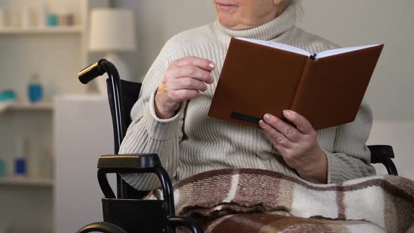 Caring Young Lady Giving Eyeglasses to Aged Granny Reading Book in Wheelchair