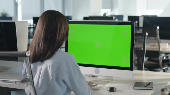 Asian Business Woman Typing on Desktop Computer with Green Mockup Screen While Sitting at His Desk