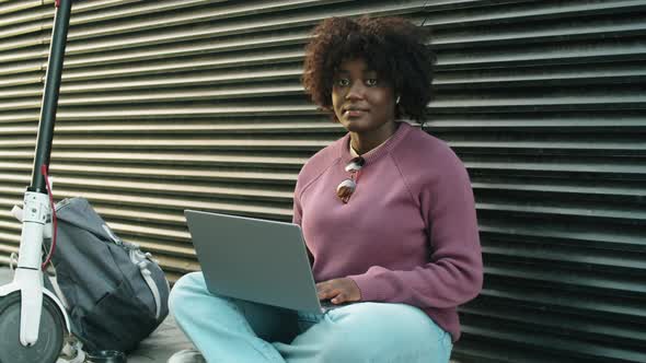 Portrait of Cheerful Afro Woman Sitting on Street and Using Laptop