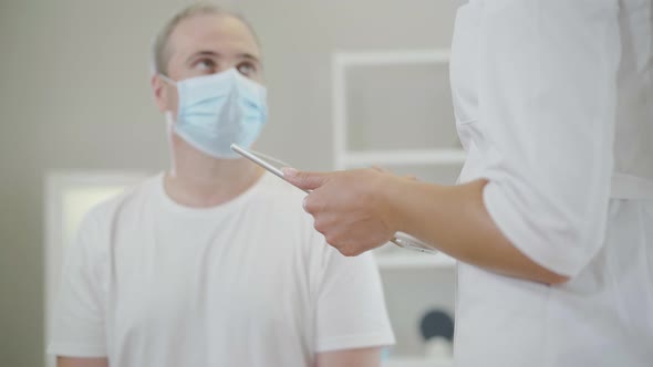Unrecognizable Doctor Taking Medical History of Mid-adult Blurred Man in Face Mask. Female Caucasian