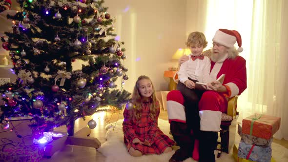Santa Clause Sitting with Little Boy and Book on Rocking Chair As Excited Girl Talking Gesturing
