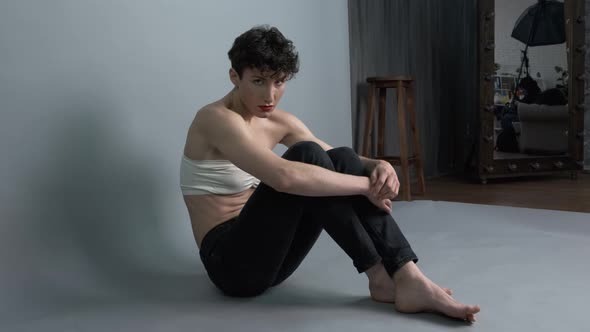 Transgender Guy with Makeup Posing While Sitting on Gray Studio Background