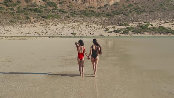 Pretty Female Friends Walking Holding Hands By Sea on Sandy Tropical Beach in Swimsuits