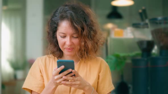 Smiling Curly Woman in Yellow Dress Using Her Phone Happily While Sits in Coffee Shop