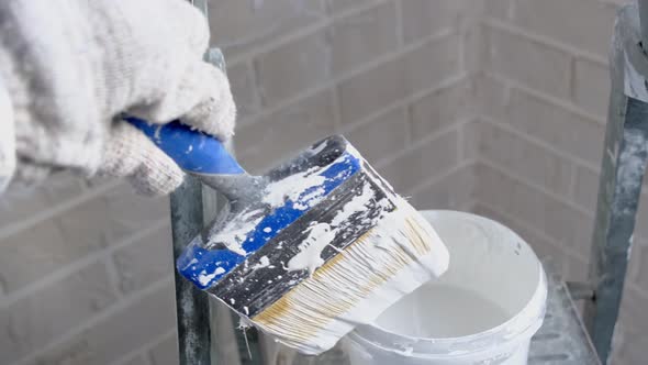 A hand in a work glove dips a paint brush in white paint in a bucket on a ladder against a brick wal