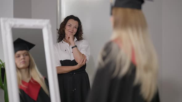 Portrait of Loving Proud Happy Mature Mother Admiring Graduate Daughter Spinning in Front of Mirror