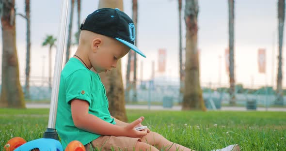 Young Boy Watches a Video on His Mobile Phone Sitting on the Grass in Park. Children and Modern